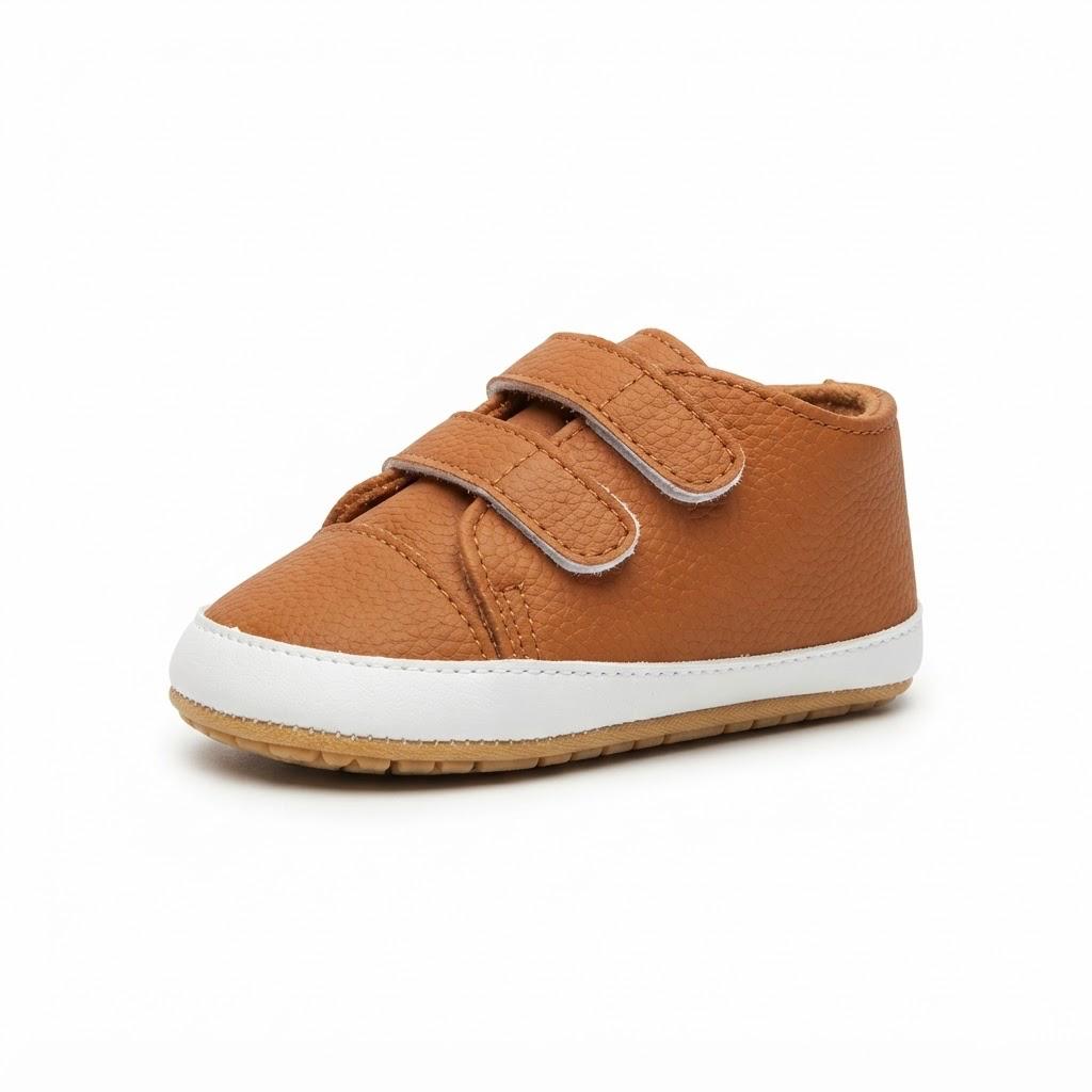A single toddler's low-top sneaker in tan pebbled leather, featuring two adjustable hook-and-loop straps with silver metal buckles, a white rubber midsole, and a flexible tan outsole, shown on a plain white background.