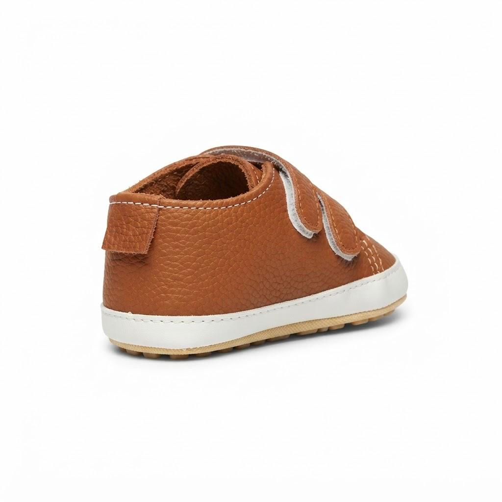 A single toddler's leather slip-on shoe in a warm brown color, featuring a textured pebbled leather upper, a white hook-and-loop strap closure, and a white rubber sole, shown from a side-rear angle on a white background.
