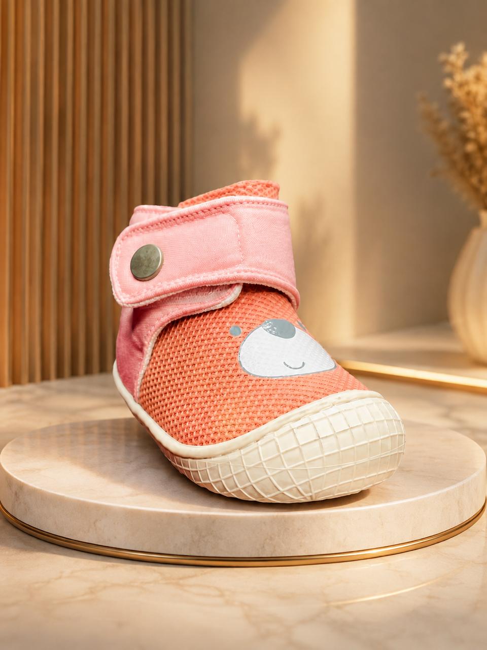 A single baby bootie in coral mesh with a pink velcro strap and a white cartoon face detail, displayed on a round marble pedestal in a warm, styled interior setting.