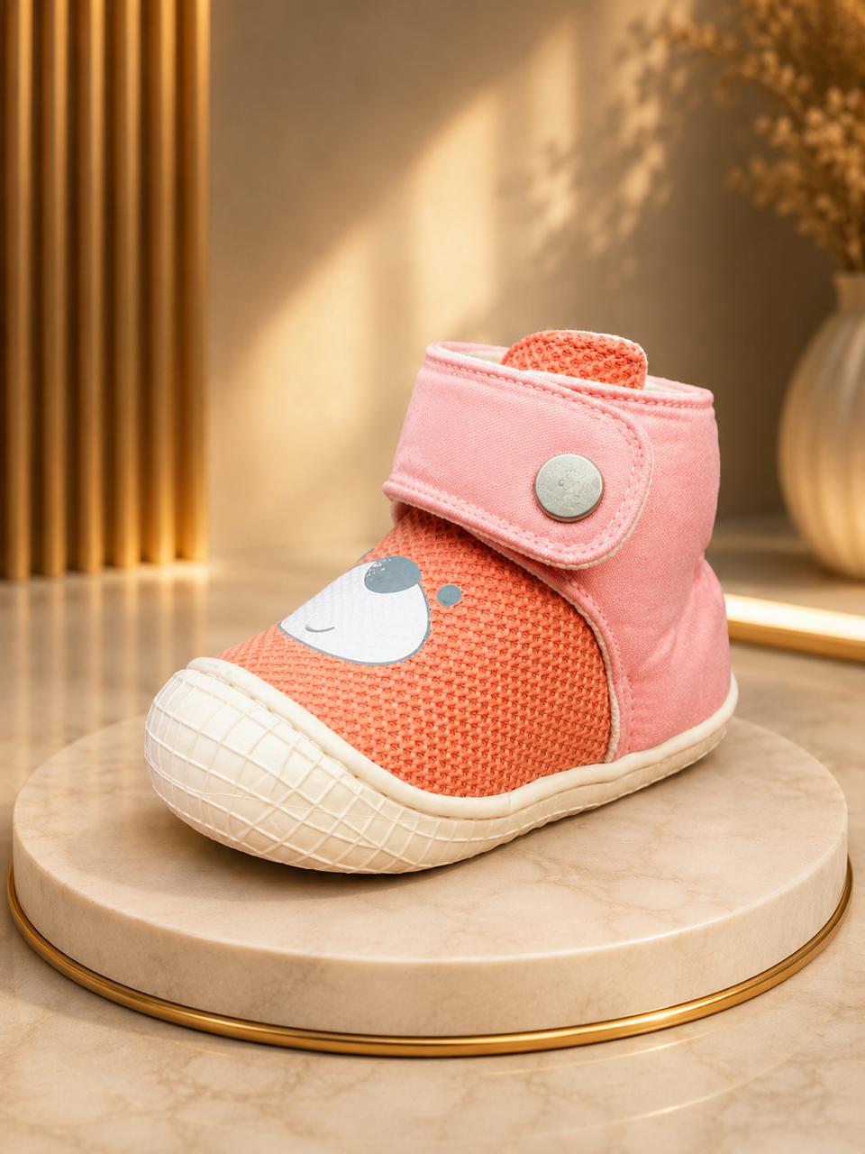 A baby bootie in coral orange mesh and pink canvas, featuring a cute cartoon face on the side, a pink strap with a silver snap, and a textured cream sole, displayed on a round marble pedestal.