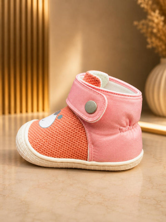 A pink baby bootie with a textured mesh side, a snap strap closure, and a small white cartoon accent on the side, resting on a beige marble surface.