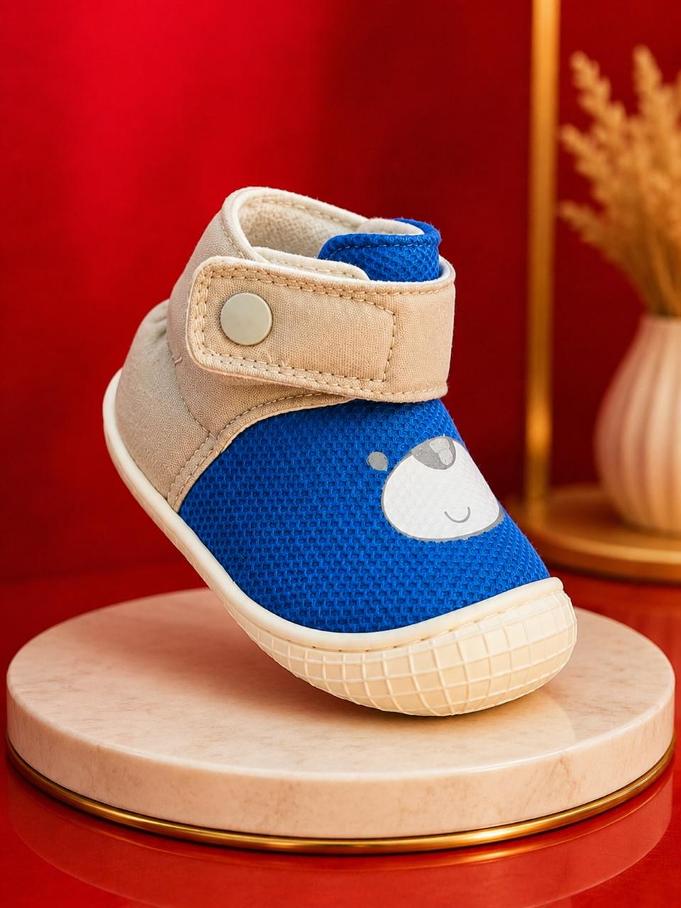 A baby's first shoe featuring a vibrant blue textured fabric upper with a white cartoon bear face, beige canvas side panels, and a beige hook-and-loop strap with a silver snap button. The shoe is displayed on a round beige marble pedestal with a gold rim against a red background.