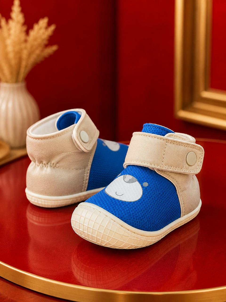 A pair of baby high-top shoes with blue mesh uppers, beige suede-like side panels, and a cream-colored sole, displayed on a glossy red surface with a decorative background.