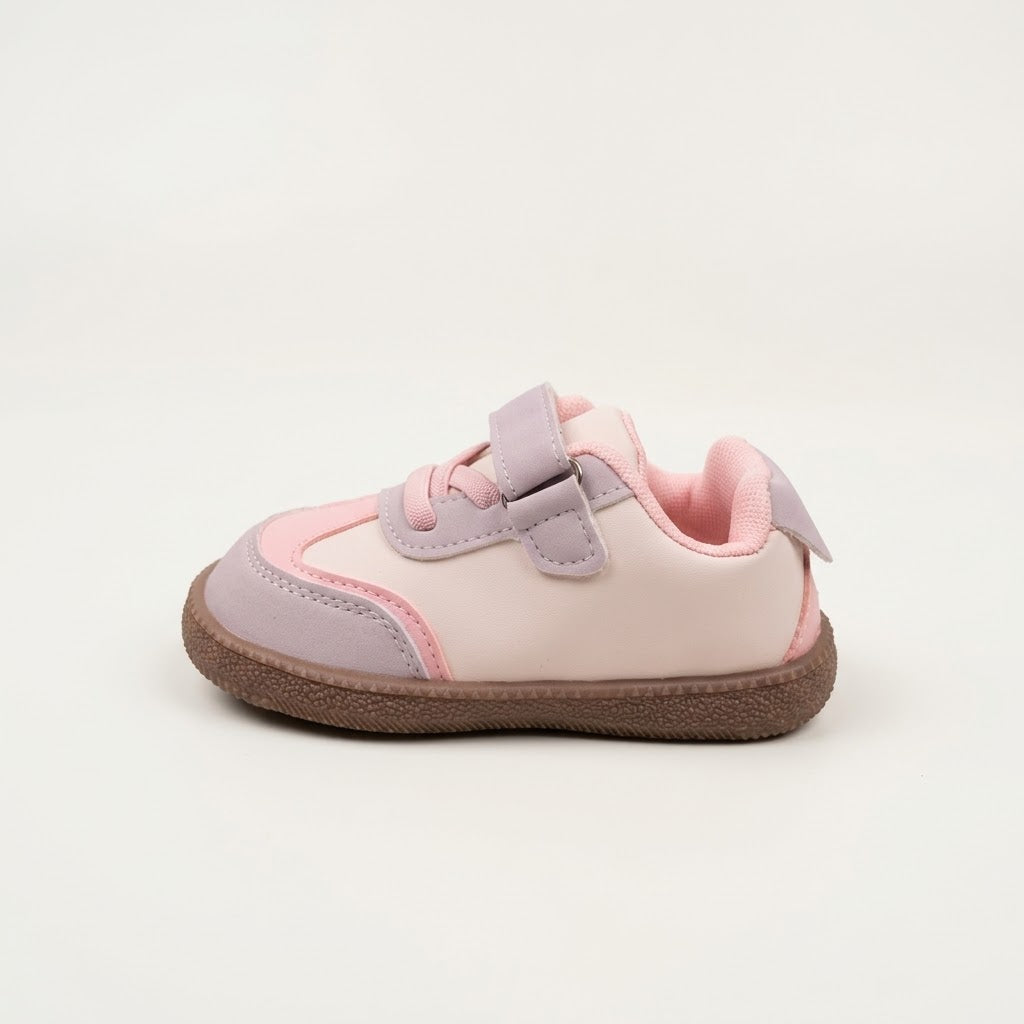 A toddler's low-top sneaker in light pink and lavender, featuring a hook-and-loop strap closure and a brown textured sole, shown against a plain light gray background.