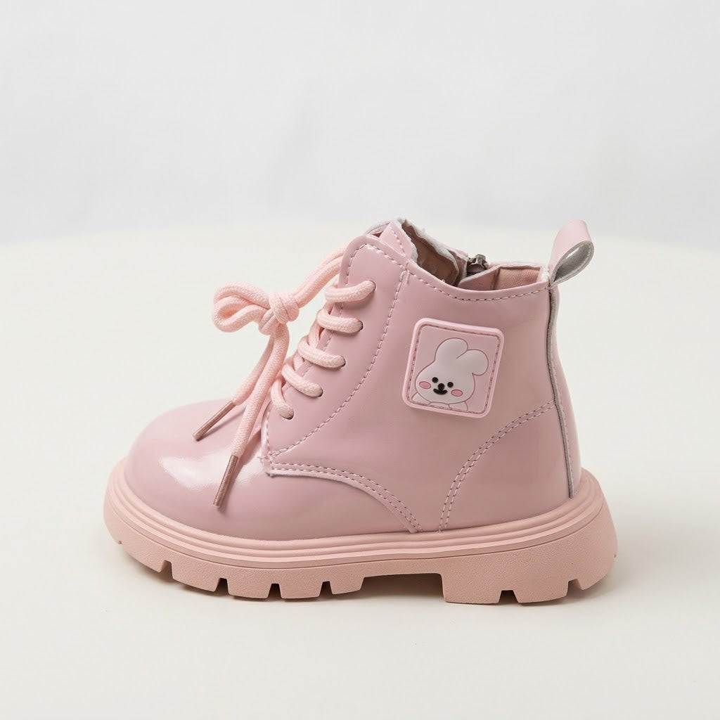 A light pink patent leather ankle boot with matching pink laces, a small cartoon patch on the side, and a chunky lug sole, shown against a plain light background.