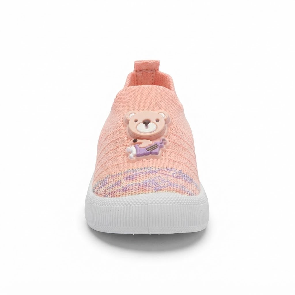 A single children's slip-on knit sneaker in a soft peach-pink color, featuring a cute 3D teddy bear applique on the side and a textured purple and pink glittery pattern near the sole, set against a plain white background.