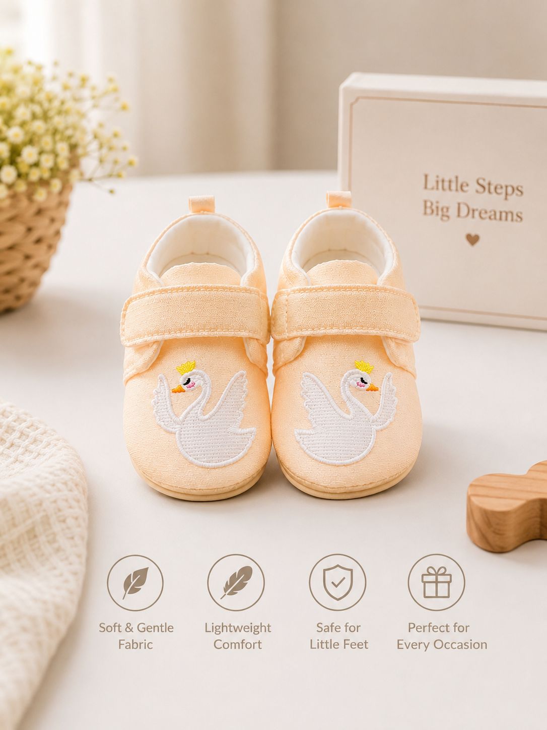 A pair of pale peach baby shoes with white swan embroidery and yellow accents, displayed on a light surface with a product box reading 'Little Steps Big Dreams' and soft decorative props in the background.