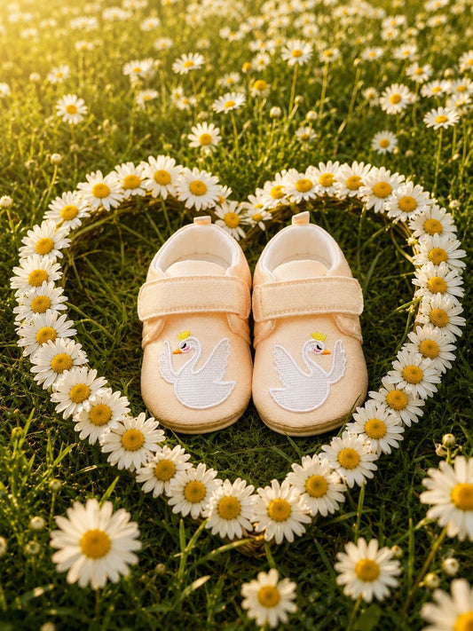 A pair of pale blush baby booties with embroidered swan designs, arranged on a grassy field surrounded by a heart-shaped border of white daisies.