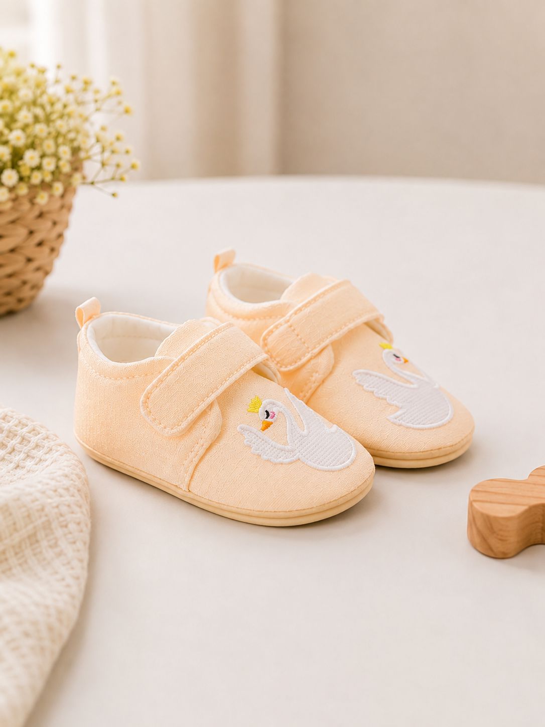 A pair of baby soft shoes in pale peach with embroidered swan designs, displayed on a light beige surface with a soft, neutral background and decorative props.
