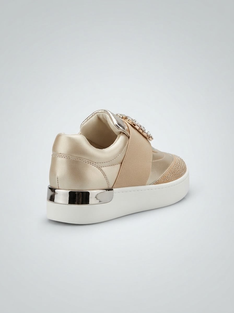 A single low-top platform sneaker in champagne beige and white, featuring a satin strap with a rhinestone-embellished buckle, a metallic silver heel accent, and a white sole, shown from a three-quarters rear angle on a light gray background.