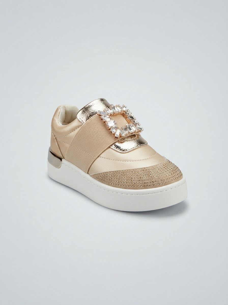 A single low-top sneaker in metallic champagne beige and gold, featuring a prominent rhinestone-embellished square buckle across the strap and a chunky white platform sole, shown on a light gray background.
